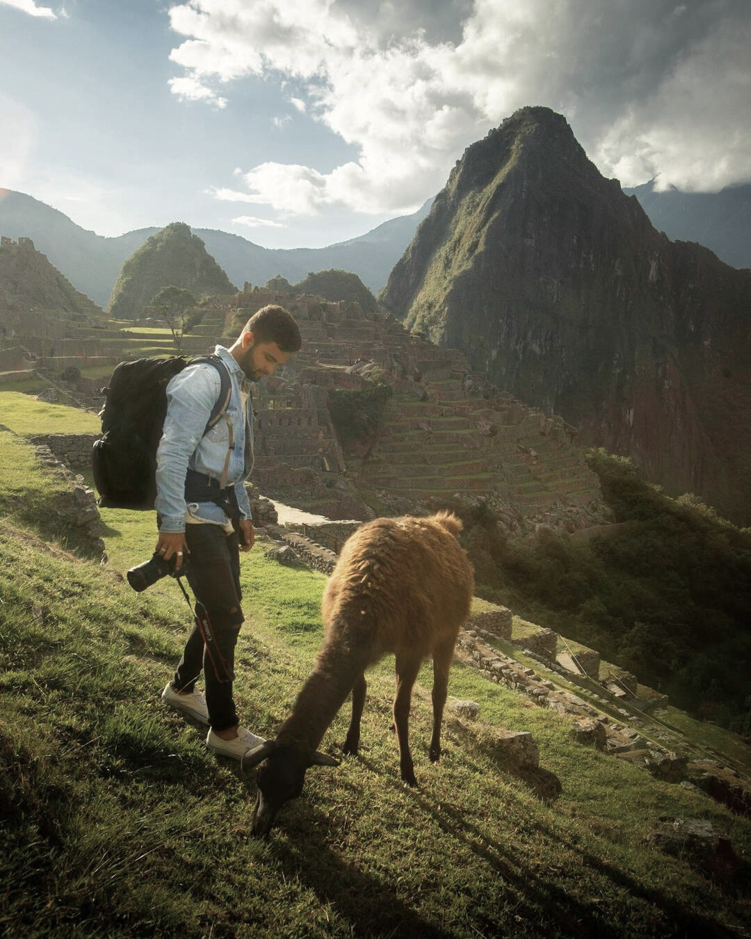 Hombre posando frente a Machu Picchu con una llama en primer plano y cámara en mano listo para fotografiar el paisaje