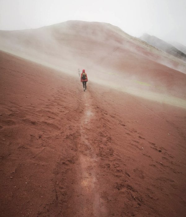 Mujer caminando por el Valle Rojo cerca de Vinicunca entre montañas rojizas y una atmósfera mística con ligera neblina en Cusco, Perú.