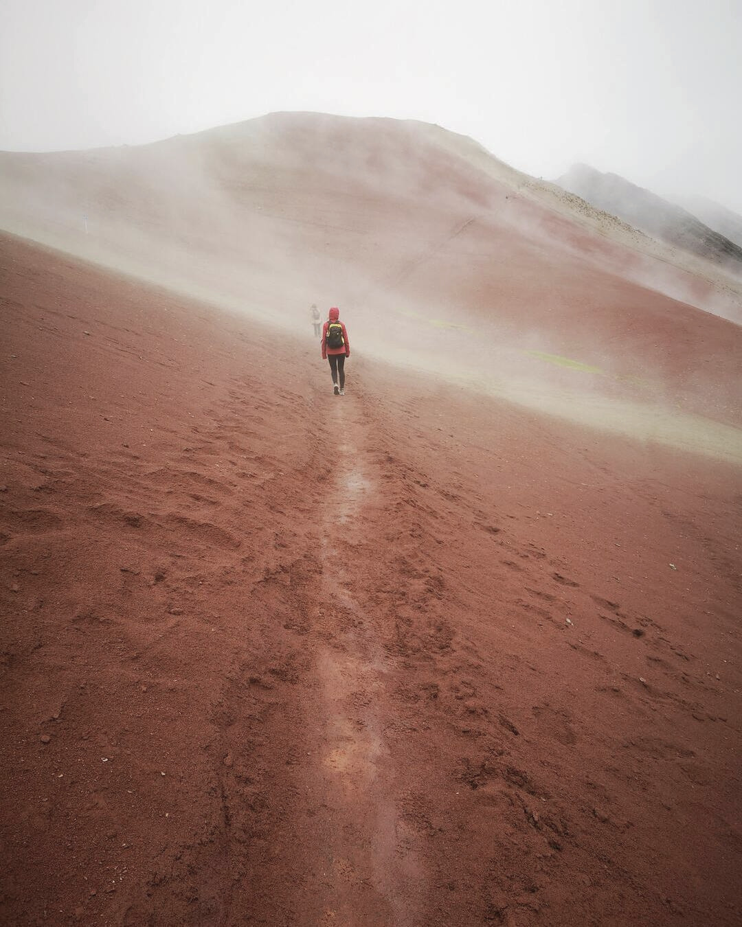 Mujer caminando por el Valle Rojo cerca de Vinicunca entre montañas rojizas y una atmósfera mística con ligera neblina en Cusco, Perú.